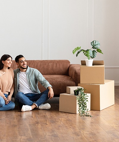 A smiling couple sits on the floor of a bright living room with a brown sofa behind them and several moving boxes with potted plants on top, suggesting they have just moved in.