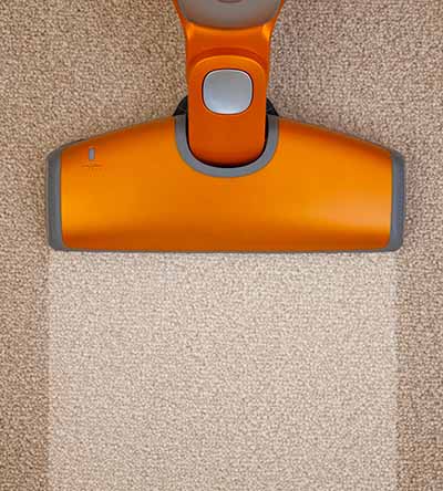 A vacuum cleaner with an orange head is cleaning a beige carpet, leaving a visible, lighter path where it has passed.