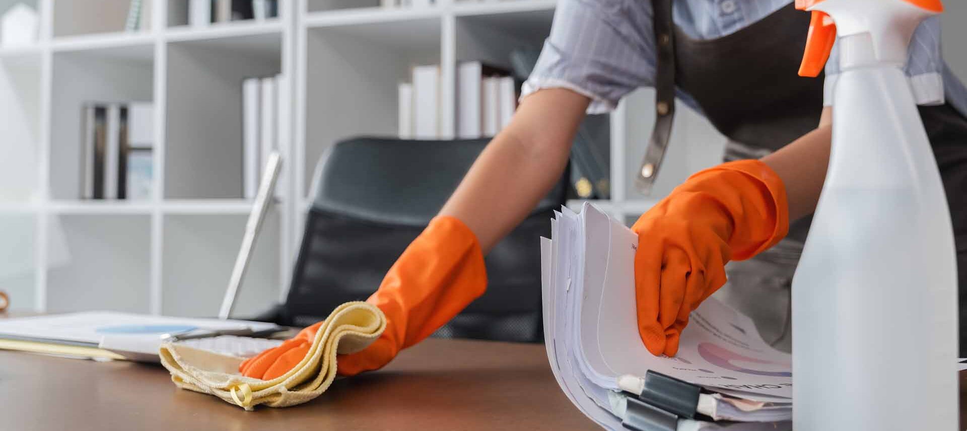 A person wearing orange gloves cleans a desk with a yellow cloth and organizes papers; cleaning spray and office shelves are visible in the background.