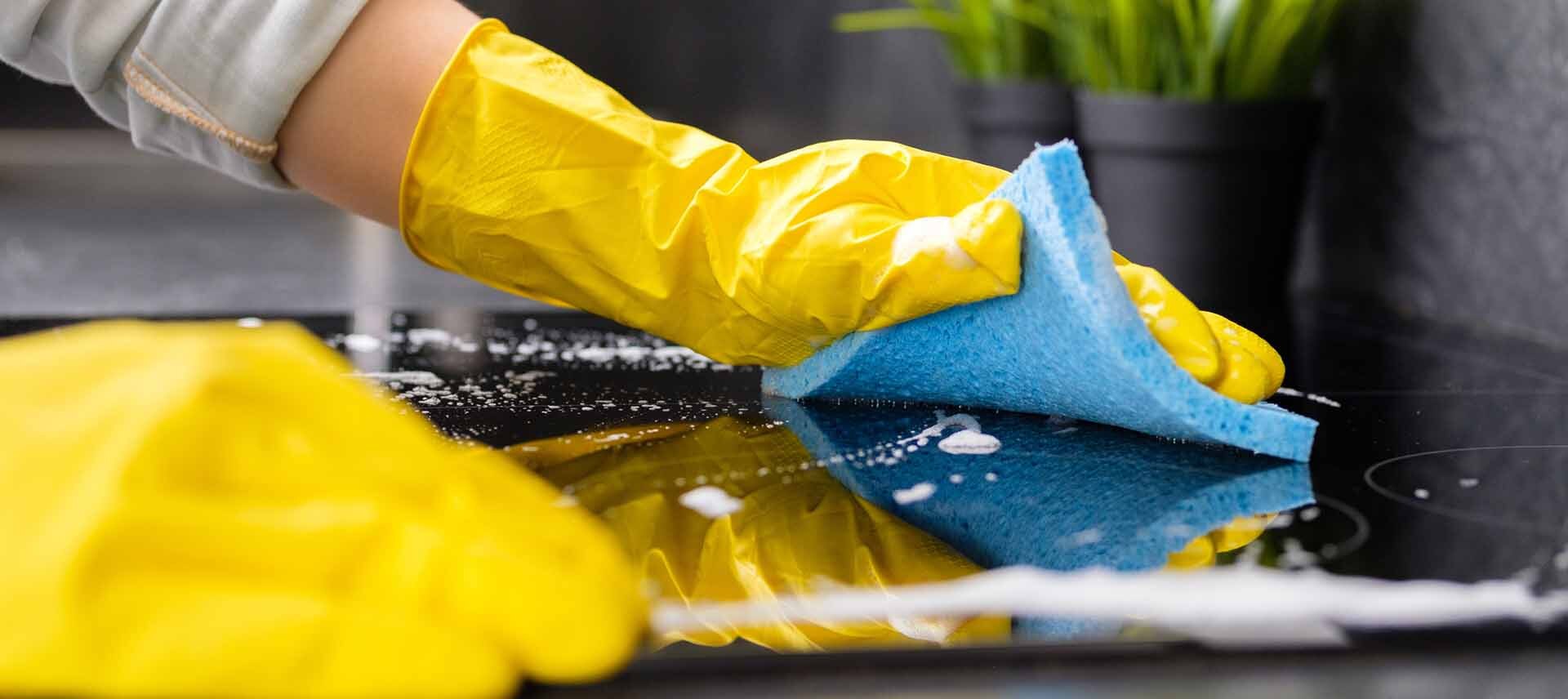 A person wearing yellow rubber gloves uses a blue sponge to clean a black glass surface with soap suds. Green potted plants are visible in the blurred background.