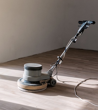 A floor buffer machine sits on a light-colored wooden floor in an empty, sunlit room with plain walls.
