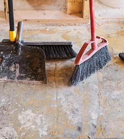 A close-up of a broom, dustpan, and push broom standing upright on a dusty, unfinished floor, suggesting a cleaning or construction site in progress.