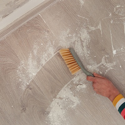 A person sweeps white dust or powder off a wooden floor with a hand brush, leaving a clean area. The person is wearing a colorful striped sleeve.