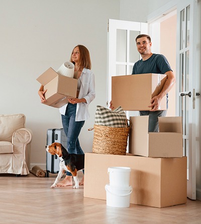 A smiling couple carrying moving boxes enters a bright living room, accompanied by a dog. More boxes, a basket, and a suitcase are placed on the wooden floor near a beige couch.