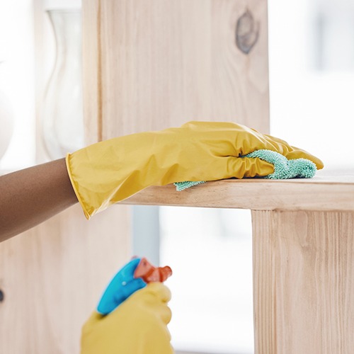 A person wearing yellow rubber gloves cleans a wooden shelf with a microfiber cloth, holding a spray bottle in the other hand.