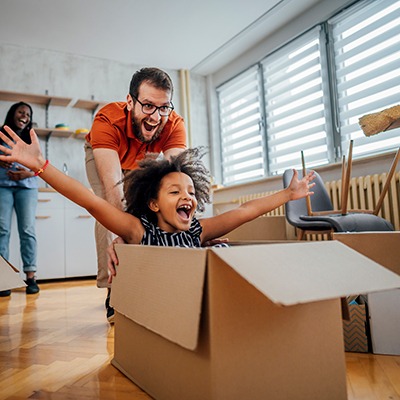 A man playfully pushes a smiling child sitting in a cardboard box, arms outstretched, inside a bright room. Another adult stands in the background, also smiling. Moving boxes and household items are visible.
