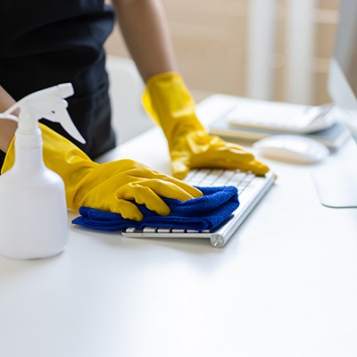 A person wearing yellow rubber gloves cleans a computer keyboard with a blue cloth. A white spray bottle sits on the white desk nearby.