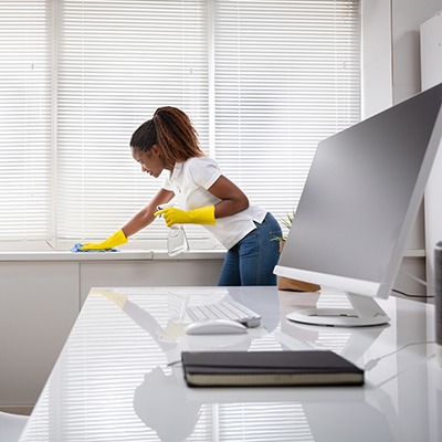 A woman wearing yellow gloves cleans a windowsill with a spray bottle and cloth in a bright, modern office with a computer, keyboard, mouse, and notebook on a white desk.