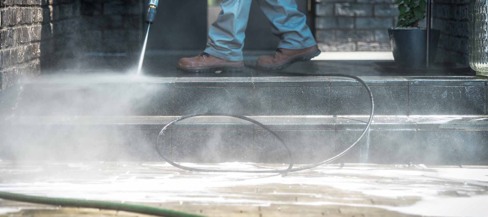 Person using a pressure washer to clean outdoor stone steps, with water spray and mist visible; a hose is coiled on the ground and a potted plant is in the background.