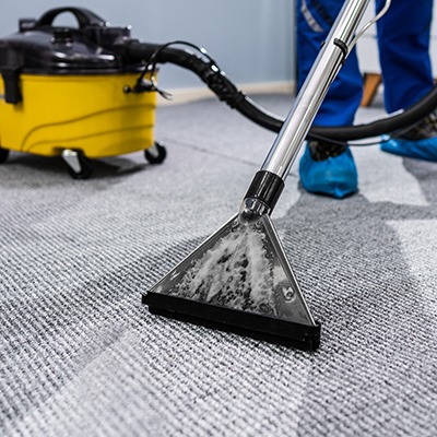 A person in blue protective shoe covers uses a carpet cleaning machine with a triangular nozzle to clean a gray carpet; a yellow vacuum unit is visible in the background.