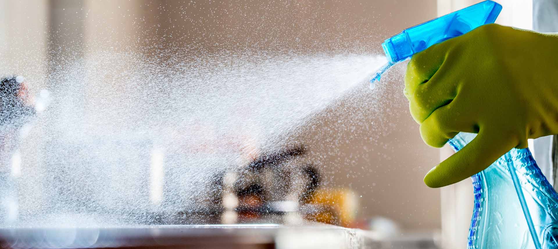 A person wearing a yellow rubber glove sprays cleaning solution from a blue spray bottle, with a fine mist spreading across the surface in a kitchen or indoor setting.