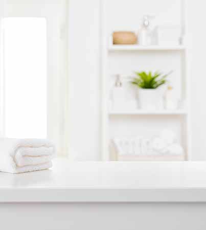 A folded white towel sits on a clean, white countertop. In the background are white shelves with neatly arranged items, including a potted green plant. The room appears bright and organized.