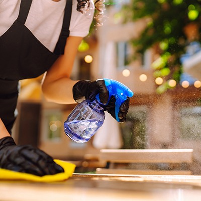 A person wearing black gloves and an apron sprays cleaner from a blue bottle onto a surface while wiping it with a yellow cloth. The background shows out-of-focus lights and greenery.