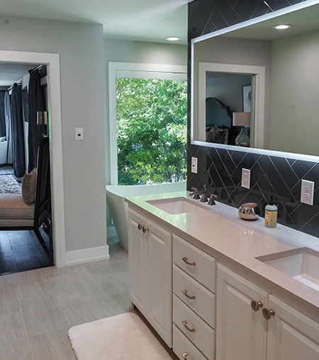 Modern bathroom with double sinks, white cabinets, a large mirror with black tile backsplash, and a freestanding bathtub by a large window; a bedroom is visible through an open doorway.