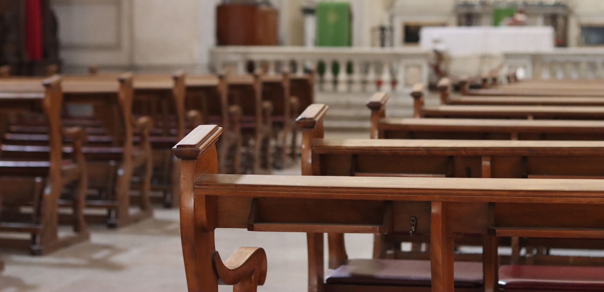 Empty wooden pews inside a church, with the altar and religious decorations visible in the background. The setting appears quiet and serene.
