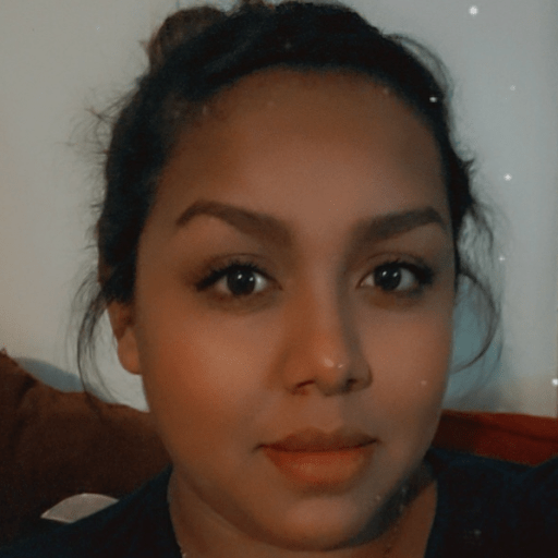 A woman with dark hair tied back looks at the camera with a neutral expression, indoors, against a plain light-colored background.