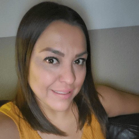 A woman with straight dark hair and a yellow sleeveless top smiles at the camera while sitting on a dark sofa. The background features a two-tone wall in light and dark shades.