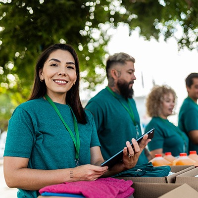 A woman in a green shirt and lanyard smiles while holding a tablet at an outdoor volunteer event. Three other people stand beside her behind tables with folded clothes and bottles of juice. Trees are visible in the background.