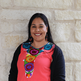 A woman with long dark hair, wearing a bright embroidered top and black cardigan, stands smiling in front of a light stone wall.