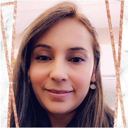 A woman with long brown hair smiles slightly at the camera. She is wearing round earrings and light makeup. The background is blurred, and a decorative border with marble and rose gold accents frames the image.