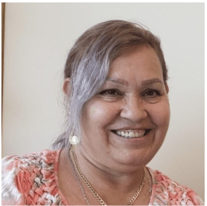 A woman with light brown hair, styled to the side, smiles warmly. She is wearing a pink and white floral top, gold necklace, and earrings, with a neutral background behind her.
