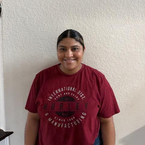 A person with dark hair tied back, wearing a red Hurley t-shirt, stands smiling in front of a plain light-colored wall.