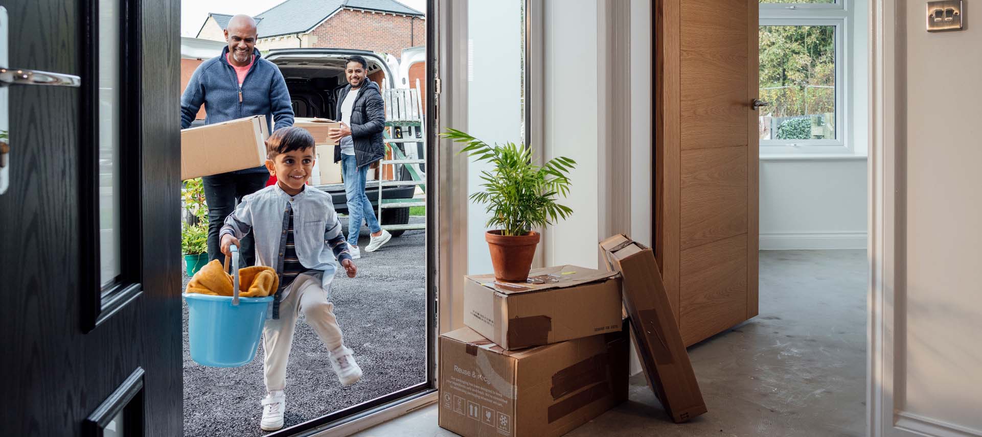 A child carrying a blue bucket enters a new home, followed by two adults with boxes. More boxes and a potted plant are stacked inside by the door, with a moving van parked outside.