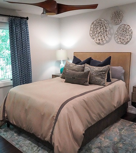 A neatly made bed with beige bedding and multiple pillows is in a cozy bedroom. A lamp sits on a bedside table, blue curtains frame the window, and decorative silver wall art hangs above the headboard.